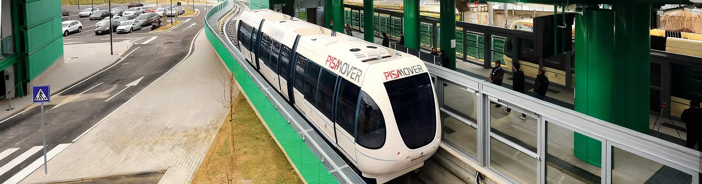 Convoglio PisaMover in arrivo alla stazione con area parcheggio sullo sfondo.